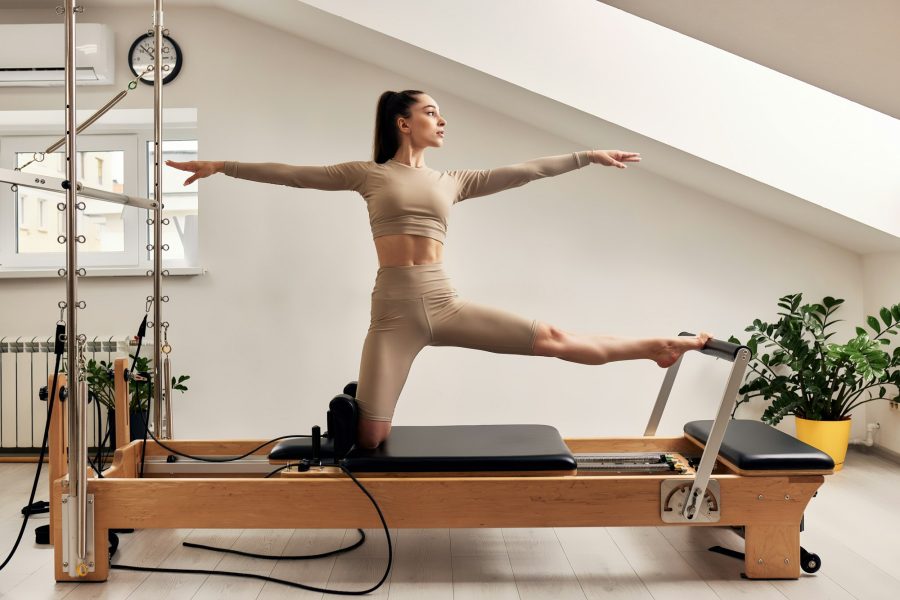 A young girl is doing Pilates on a reformer bed. A beautiful slender brunette woman in a beige