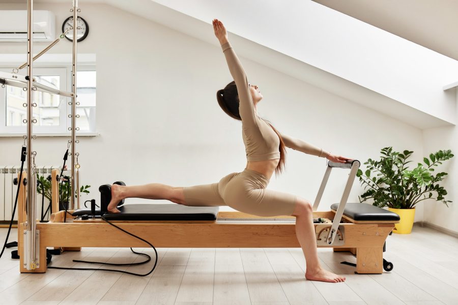 A young girl is doing Pilates on a reformer bed in a bright studio. A beautiful slender brunette in