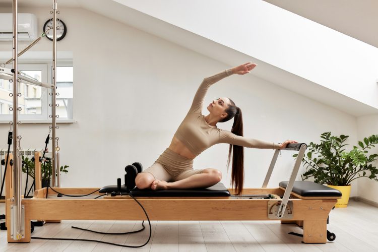 A young girl is doing Pilates on a reformer bed in a bright studio. A slender brunette in a beige